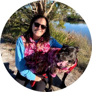 Woman with brown hair wearing sunglasses, long sleeves, and a matching pink sweater with a black dog with their tongue hanging out in a coastal Florida setting. 