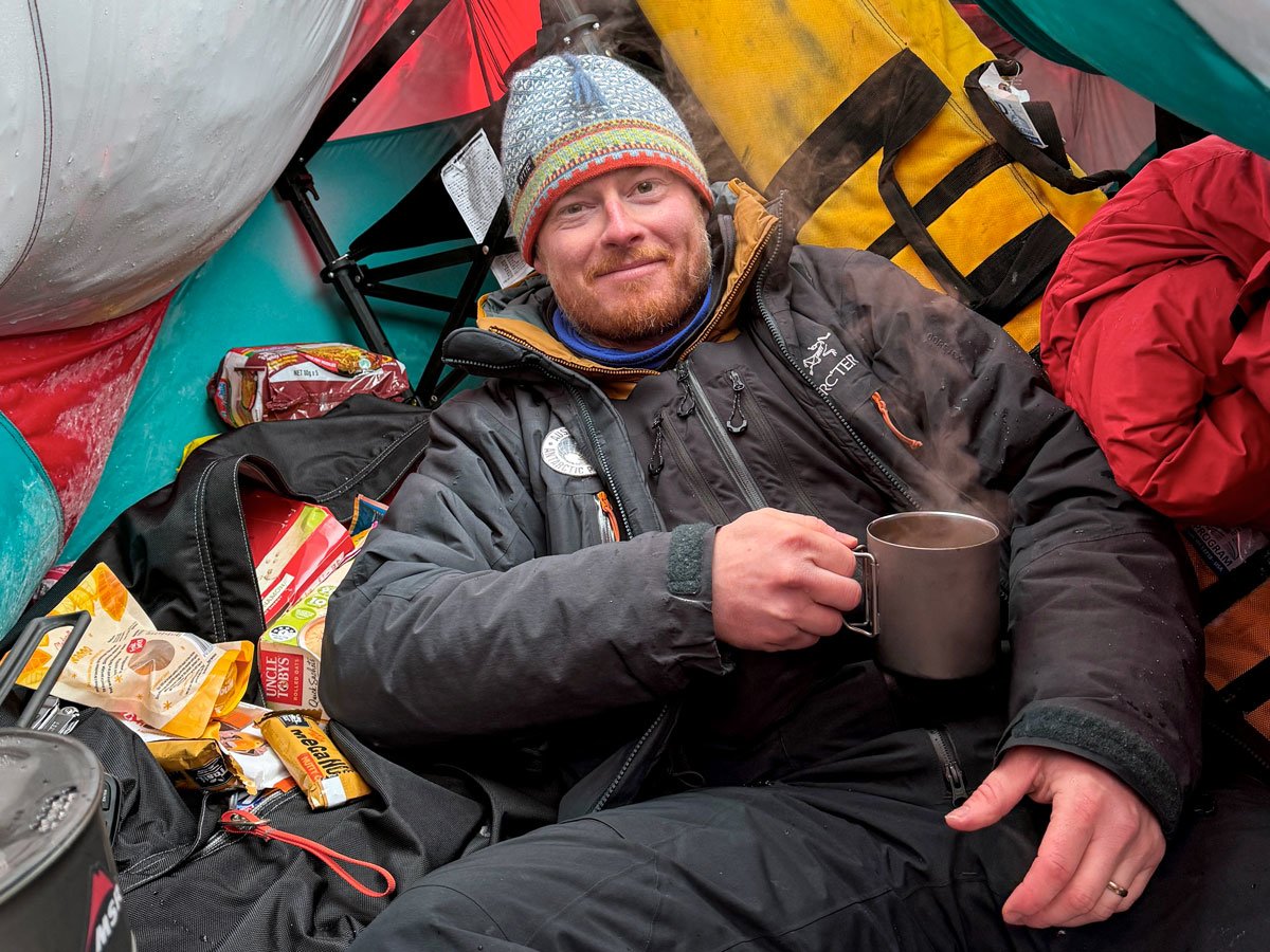 man in winter gear in tent holding mug of steaming coffee